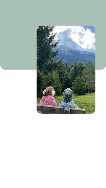 liebevoll aufwachsen – Zwei Kinder sitzen auf einer Bank auf einer grünen Wiese, schauen in die Ferne auf bewaldete Hügel und Berge, umgeben von Natur und Stille.