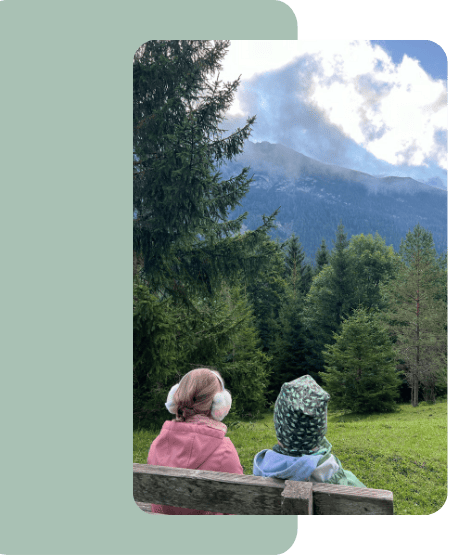 liebevoll aufwachsen – Zwei Kinder sitzen auf einer Holzbank mit Blick auf einen bewaldeten Hang und Berge – ein stiller Moment in der Natur, voller Ruhe und Verbundenheit.