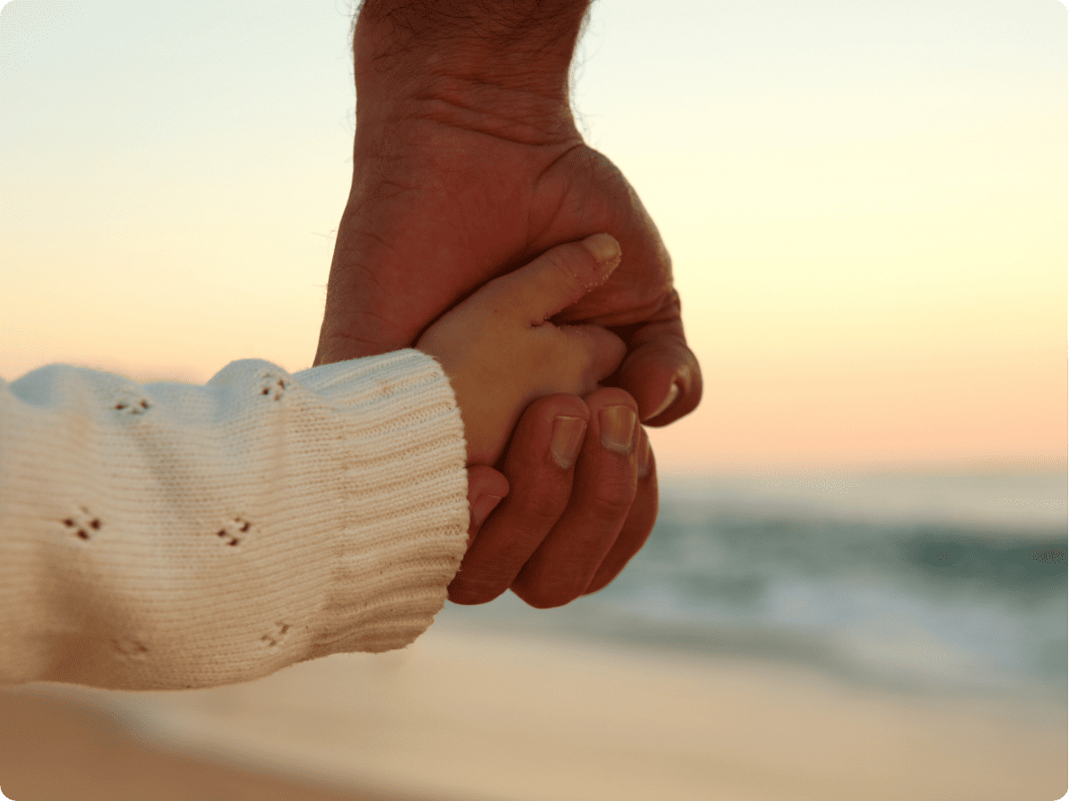 liebevoll aufwachsen - Ein Erwachsener hält die Hand eines Kindes bei Sonnenuntergang am Strand als Symbol für Geborgenheit und Bindung